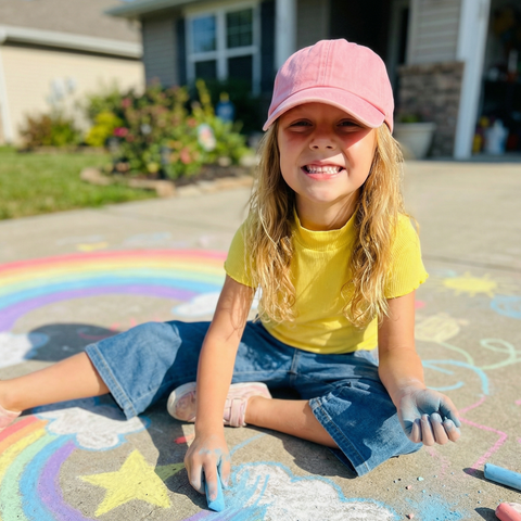 A young girl with long blonde hair sits on a painted rainbow chalkboard on a concrete driveway, wearing a pink baseball cap, yellow t-shirt, and denim shorts, with her hands covered in chalk.