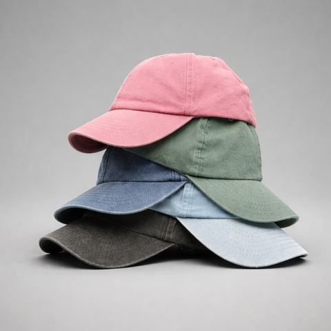 Four fabric baseball caps stacked together on a neutral gray background, showing different muted color options: pink, sage green, dusty blue, and charcoal gray.