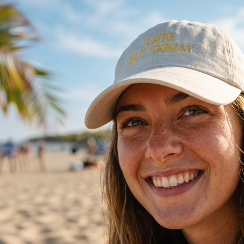 A smiling woman on a beach wearing a light beige baseball cap with yellow embroidered text that reads 'LATE BUT GREAT'.