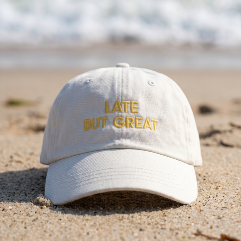 A white cotton baseball cap with 'LATE BUT GREAT' embroidered in yellow on the front, resting on a sandy beach with the ocean blurred in the background.