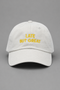 A white cotton baseball cap with yellow embroidered text reading 'LATE BUT GREAT' on the front, photographed against a solid gray background.