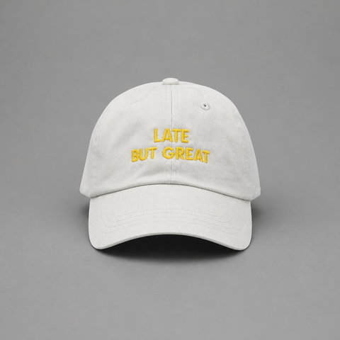 A white cotton baseball cap with yellow embroidered text reading 'LATE BUT GREAT' on the front, photographed against a solid gray background.