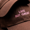 A close-up of a dark brown baseball cap with pink embroidered text reading 'LATE BUT GREAT' on the front panel.