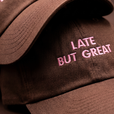 A close-up of a dark brown baseball cap with pink embroidered text reading 'LATE BUT GREAT' on the front panel.