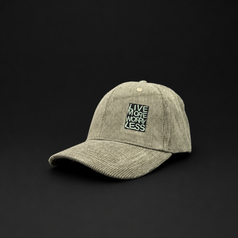 A light olive green corduroy baseball cap with a black embroidered square patch on the front that reads 'LIVE MORE WORRY LESS'. The cap is photographed on a solid black background.