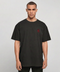 A man wearing an oversized black t-shirt with a small red embroidered skull graphic on the left chest, photographed against a neutral light gray background.