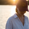A woman outdoors by the water at sunset wearing a burgundy baseball cap with gold embroidered text that reads 'be bold or italic, but never regular' and a light blue button-up shirt.