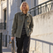 A woman stands outdoors on a city sidewalk holding a skateboard, wearing an olive green button-up utility jacket over a black and white striped shirt with dark pants.