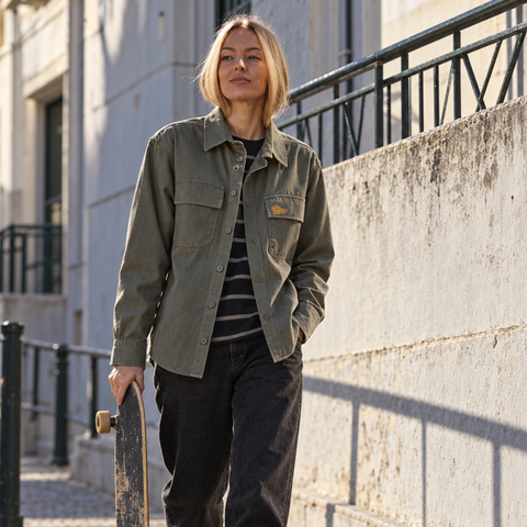 A woman stands outdoors on a city sidewalk holding a skateboard, wearing an olive green button-up utility jacket over a black and white striped shirt with dark pants.