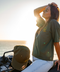 A woman sits on a motorcycle by the ocean at sunset, wearing an olive green t-shirt with a small gold chest logo and a matching olive green baseball cap resting on the motorcycle. The background shows the bright sun over the water.
