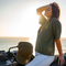 A woman sits on a motorcycle by the ocean at sunset, wearing an olive green t-shirt with a small gold chest logo and a matching olive green baseball cap resting on the motorcycle. The background shows the bright sun over the water.