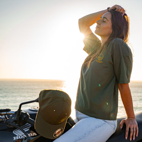A woman sits on a motorcycle by the ocean at sunset, wearing an olive green t-shirt with a small gold chest logo and a matching olive green baseball cap resting on the motorcycle. The background shows the bright sun over the water.