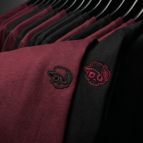 Close-up of a row of crewneck sweatshirts on black hangers, showing a heathered burgundy/maroon version with a black embroidered skull wearing a hat, and a black version with a red embroidered skull wearing a hat.