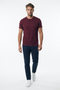A man wearing a short-sleeve crewneck t-shirt in maroon, paired with dark navy pants and white sneakers, walking forward against a solid white studio background.