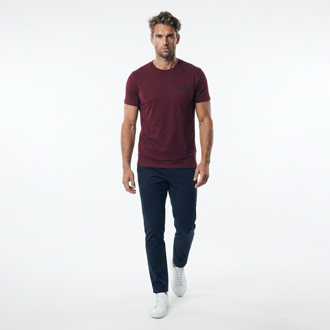 A man wearing a short-sleeve crewneck t-shirt in maroon, paired with dark navy pants and white sneakers, walking forward against a solid white studio background.