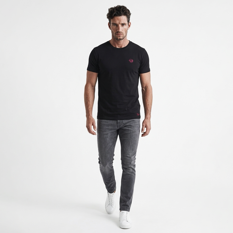 A man modeling a black short-sleeve crewneck t-shirt with a small red embroidered logo on the left chest, paired with gray jeans and white sneakers, against a plain white studio background.