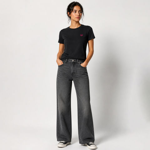 A woman modeling a black short-sleeve t-shirt with a small pink embroidered logo on the left chest, paired with dark gray wide-leg jeans and black-and-white sneakers, against a light gray studio background.