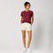 A woman modeling a short-sleeve crewneck t-shirt in burgundy with a small black embroidered logo on the left chest, paired with off-white shorts and white sneakers, against a light gray studio background.